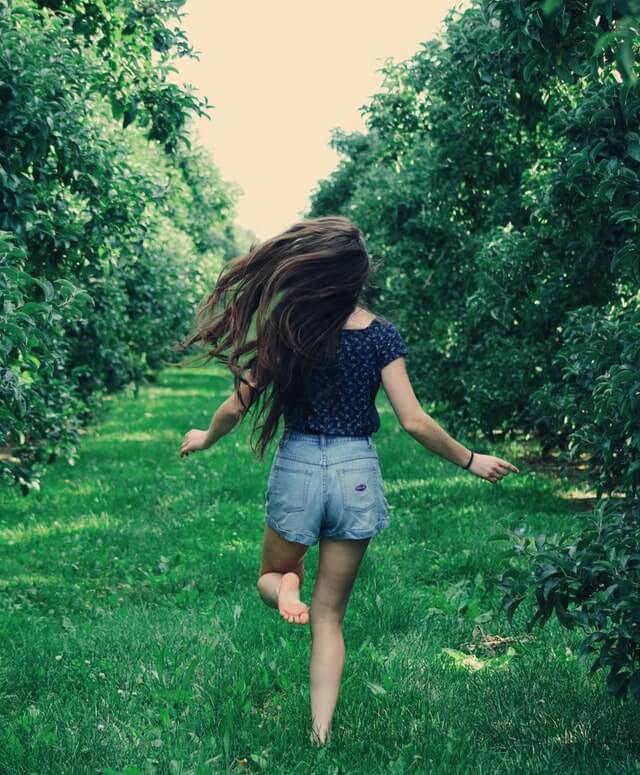 En ung pige i sommertøj, der løber i bare tæer på græs, omringet af grønne træer